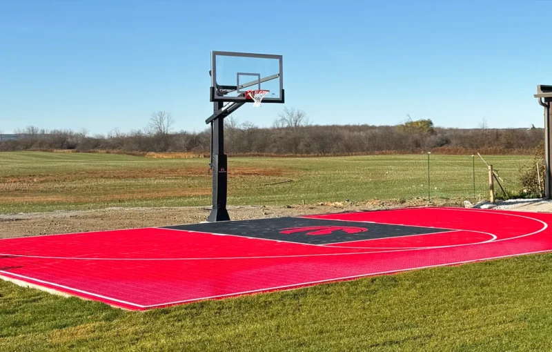 A custom design Toronto Raptors red and black backyard basketball court installed by Elite Courts next to a field