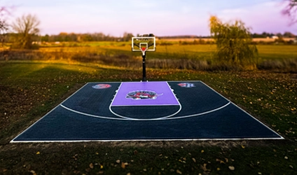 A custom design Toronto Raptors purple and black backyard basketball court installed by Elite Courts next to a field
