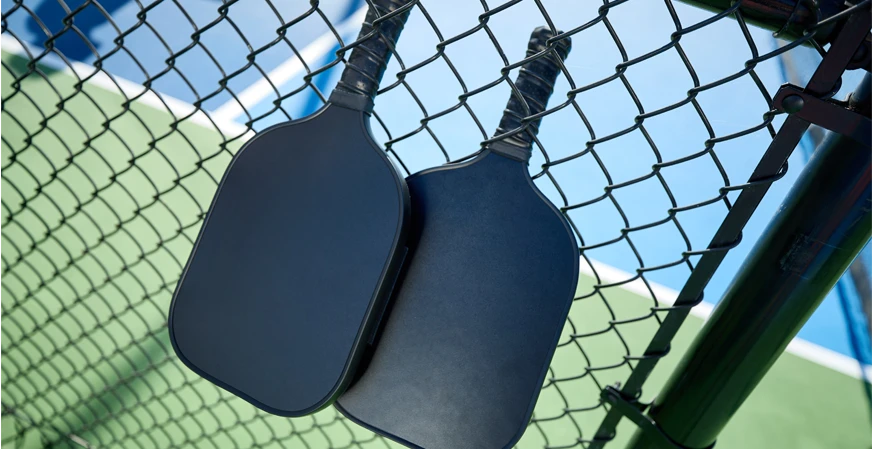 Two black pickleball paddles on a black fence next to the pickleball court